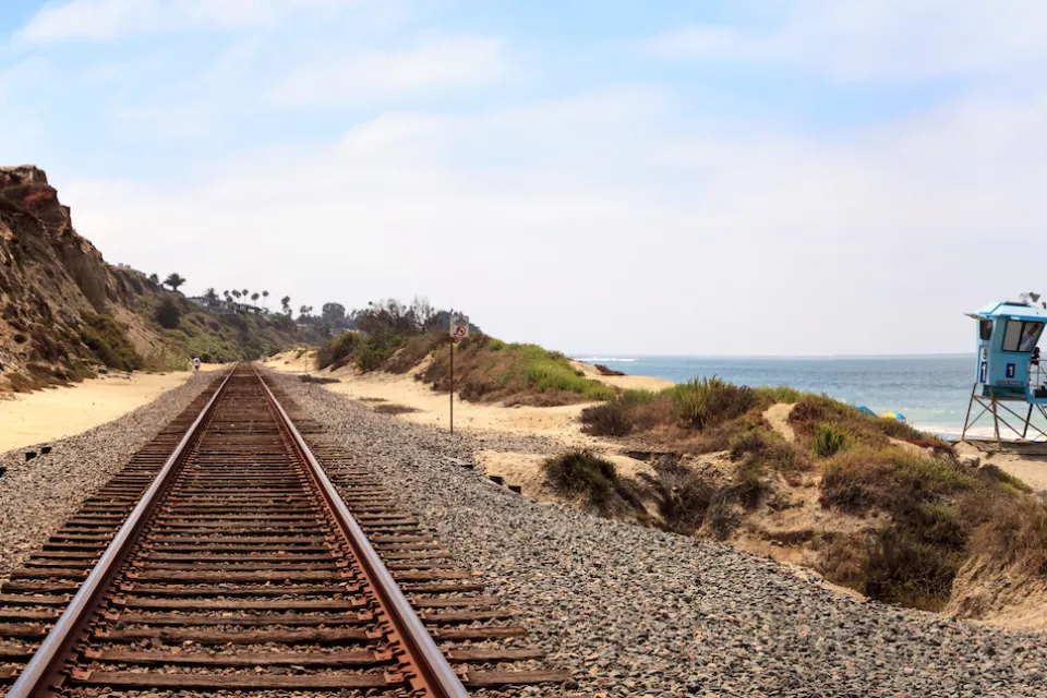 View looking down train tracks at San Clemente, California with grassy beach dunes and blue lifeguard tower on right side.